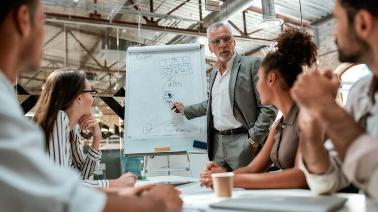 An experienced professional leads a diverse team in a collaborative discussion, explaining concepts on a flipchart, emphasizing the theme of 'Leadership Across Cultures'